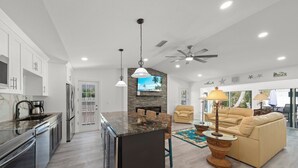 A modern kitchen with a central island, a dining area with a sofa, and a living room with a ceiling fan. - Tipsy Turtle: Charming 3-Bedroom Beach Cottage! (Sanibel)