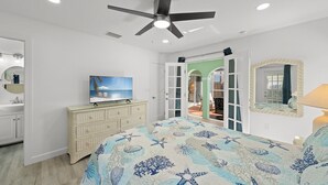 A bedroom with a bed, a ceiling fan, a television, a dresser, a mirror, and a view of an outdoor patio. - Tipsy Turtle: Charming 3-Bedroom Beach Cottage! (Sanibel)