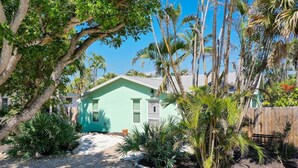 A small, single-story house with a green exterior, surrounded by tropical vegetation and a gravel driveway. - Tipsy Turtle: Charming 3-Bedroom Beach Cottage! (Sanibel)