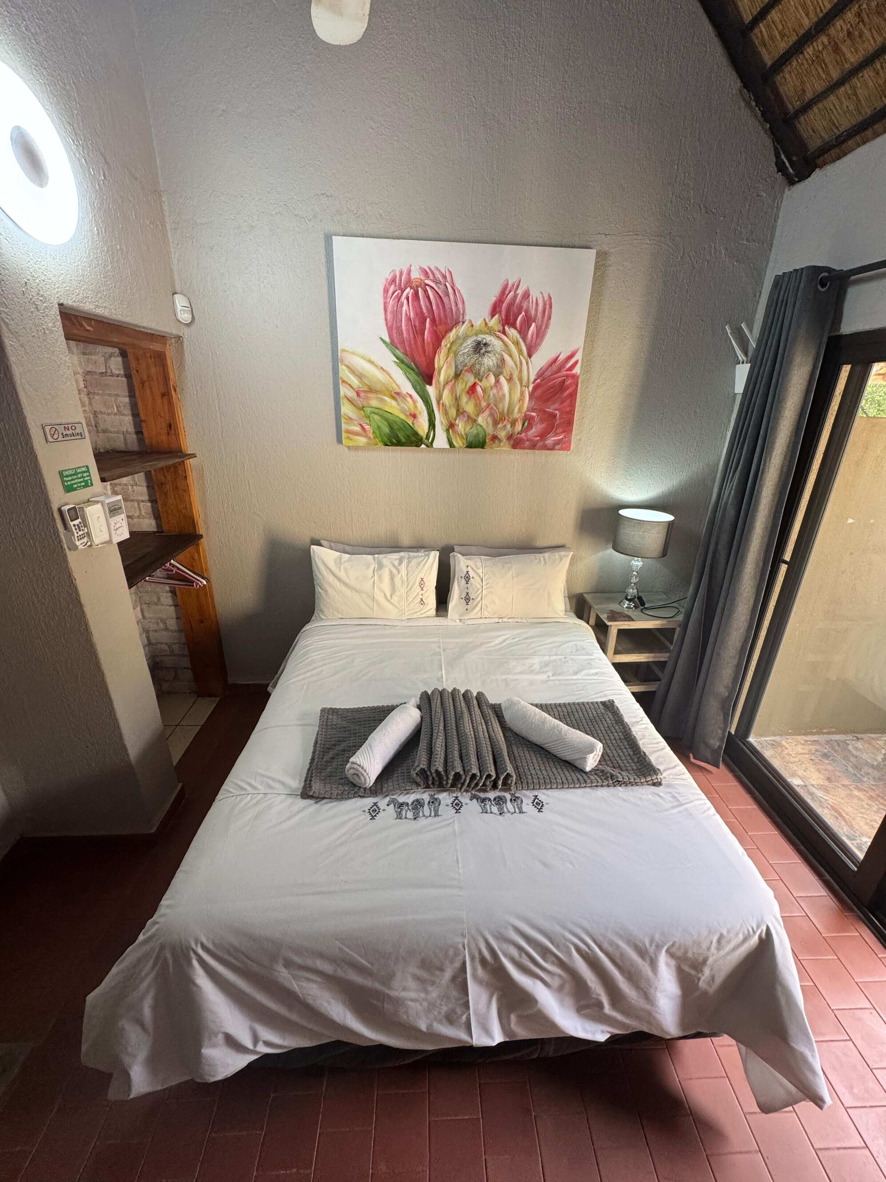 Een net opgemaakt bed met een witte dekbedovertrek en kussens, een bloemschilderij aan de muur, een tafeltje met een lamp en een raam met gordijnen.