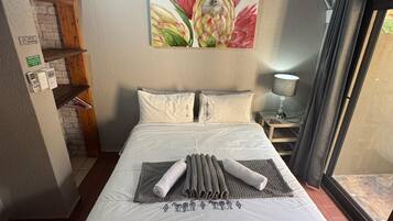 Een net opgemaakt bed met een witte dekbedovertrek en kussens, een bloemschilderij aan de muur, een tafeltje met een lamp en een raam met gordijnen.