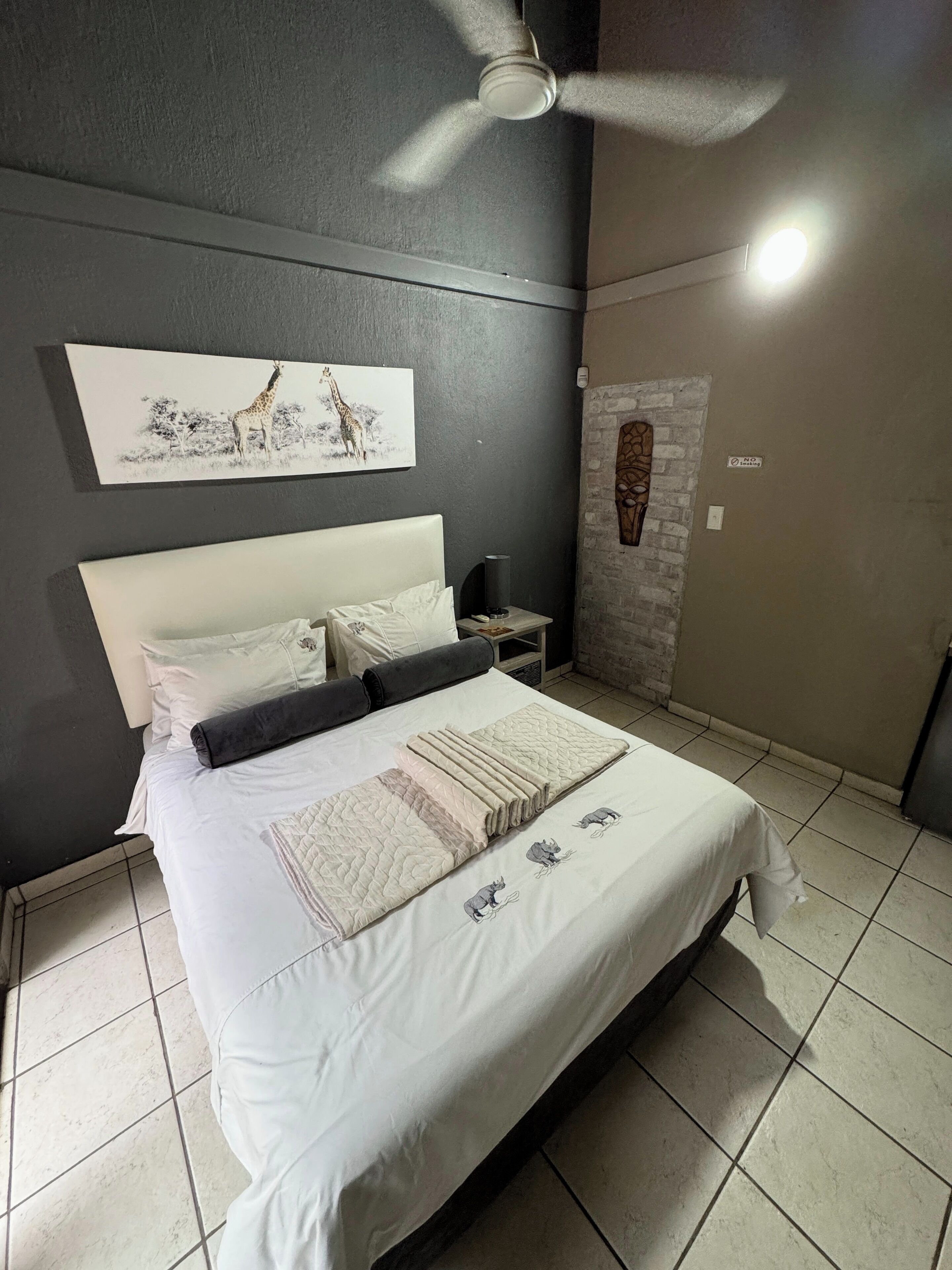 A neatly made bed with a headboard, a bedside table with a lamp, a wall-mounted artwork of giraffes, and a ceiling fan.
