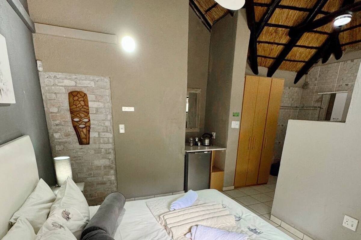 Un dormitorio con cama, almohadas, una mesita de noche, una lámpara, un ventilador de techo y una zona de cocina al fondo.