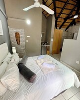 A bedroom with a bed, pillows, a bedside table, a lamp, a ceiling fan, and a kitchen area in the background.