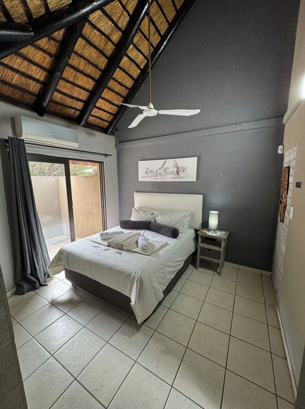 Una camera da letto con un letto, un comodino, una ventola a soffitto, una finestra con tende e un'opera d'arte appesa al muro.