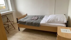 Una cama individual con una colcha gris y almohadas blancas, una silla de madera y un radiador debajo de una ventana.