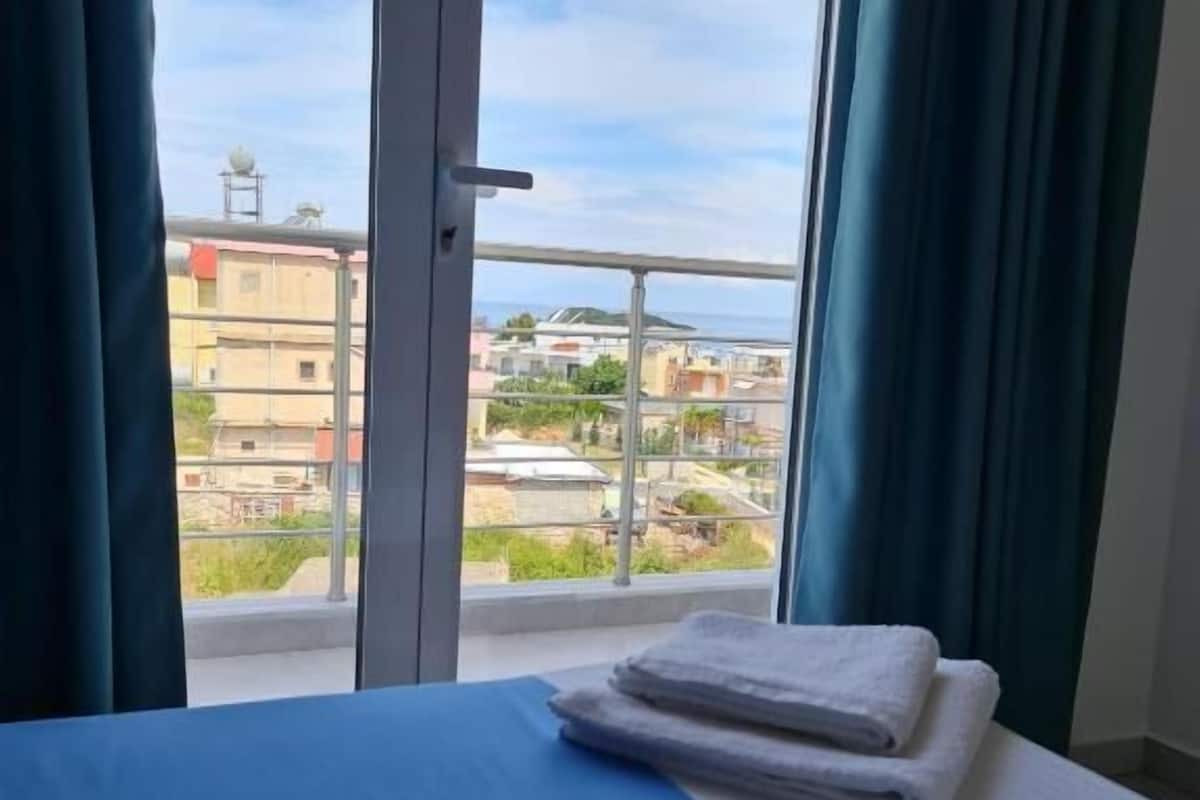 Habitación de hotel con cama, vistas a edificios y vegetación, y un balcón con barandilla.