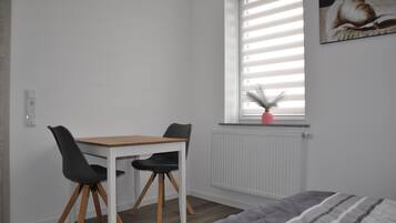 Ein kleiner, ordentlich eingerichteter Raum mit einem weißen Tisch, zwei schwarzen Stühlen, einem Fenster mit Jalousien, einer Heizung und einem Bett mit einer gestreiften Decke.