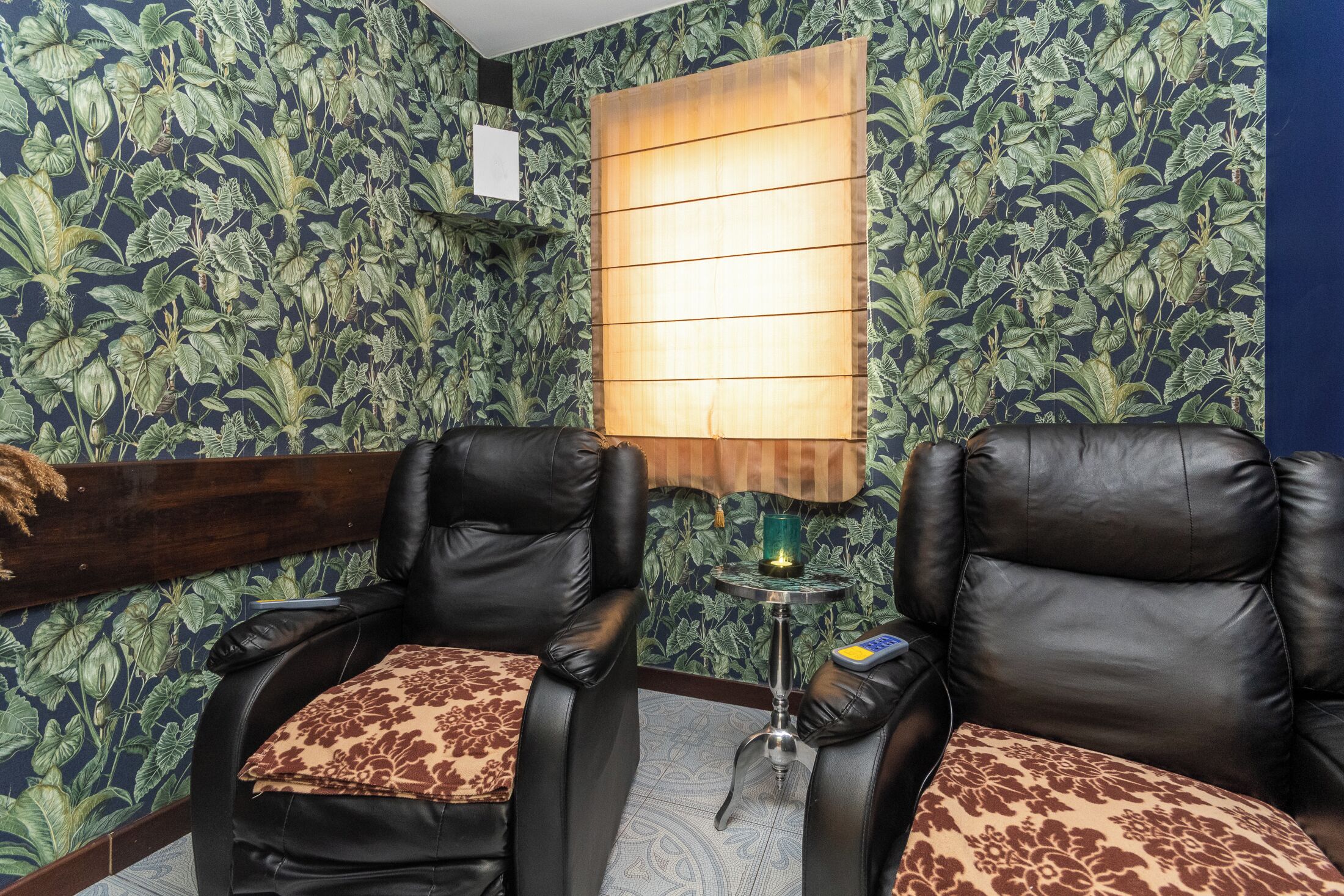 A room with two leather chairs, a small table with a lamp, and a wall with a leafy pattern.