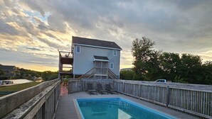 A rectangular swimming pool with a wooden deck in the foreground, a wooden fence surrounding the pool, and a two-story house with a balcony in the background. - 5-bedroom house in tranquil Corolla North Carolina OBX 4x4 Carova (Corolla)