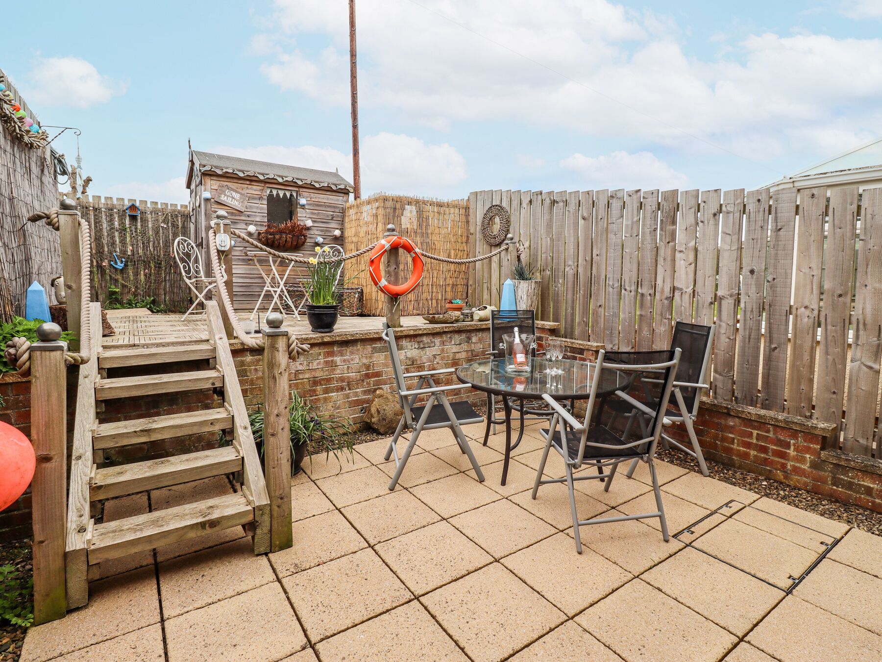 A patio with a glass table, metal chairs, a wooden bench, a small staircase, and a wooden fence.