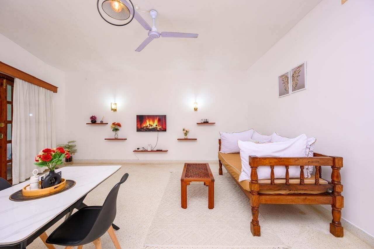 Una habitación con una cama de madera, una mesa de comedor con sillas, un taburete de madera pequeño, un ventilador de techo y estantes en la pared con adornos.