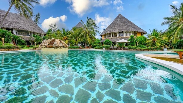 Een resort zwembad met helder water, omgeven door palmbomen en gebouwen met rieten daken.