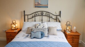 A neatly made bed with a blue quilt, multiple pillows, two bedside tables with lanterns, and a painting of a lighthouse on the wall. - Tee View (Seahouses)