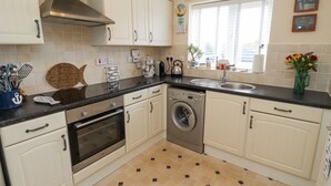 A compact kitchen with white cabinets, a stainless steel range hood, a built-in washing machine, and a window with blinds. - Tee View (Seahouses)
