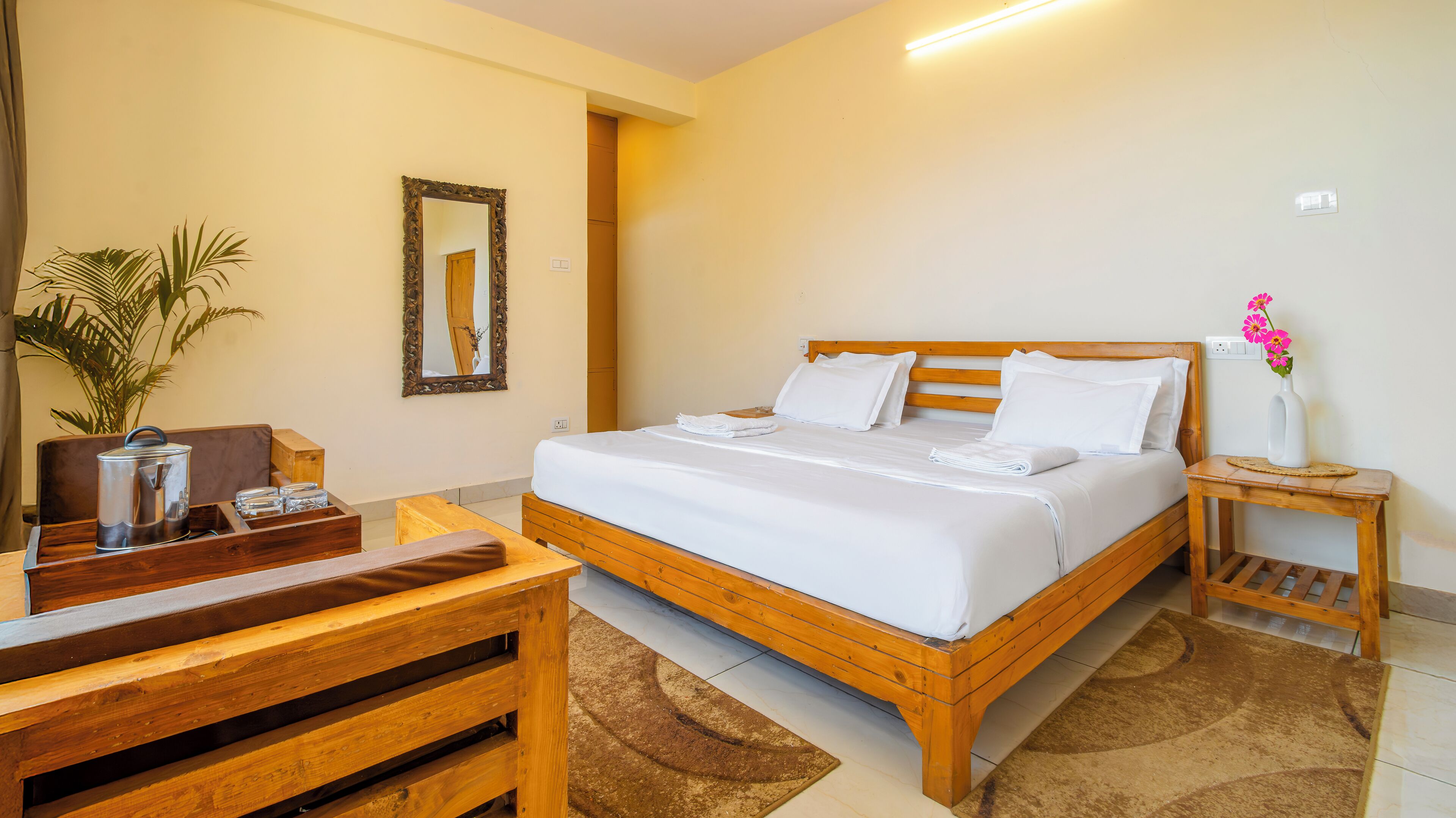 Ein ordentlich eingerichtetes Hotelzimmer mit einem Holzbett, einem Nachttisch mit Blumenvase, einem Holztisch mit Wasserkocher und einem Spiegel an der Wand.