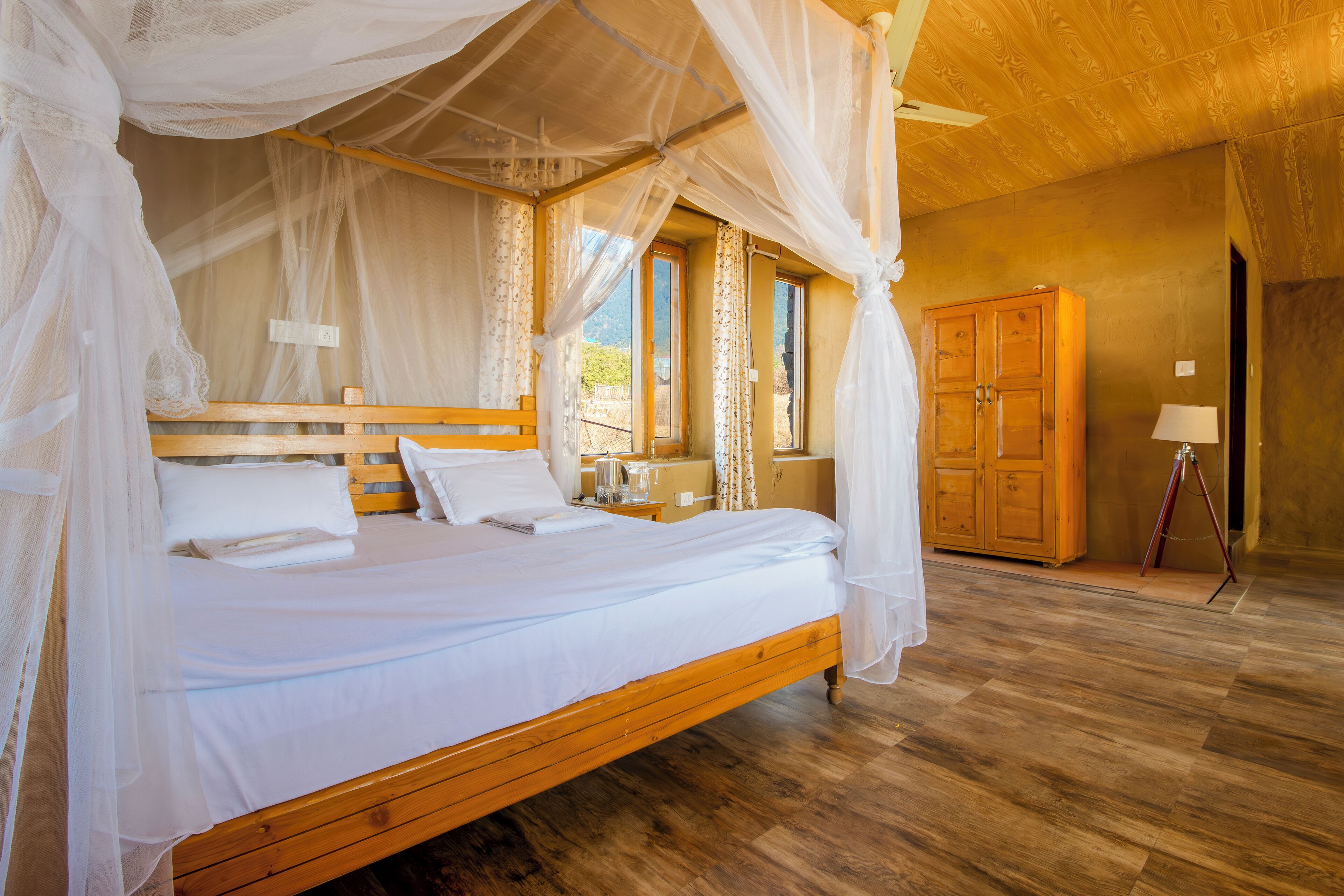 A wooden four-poster bed with white linens, a wooden nightstand, and a wooden wardrobe in a room with wooden flooring and a wooden ceiling.