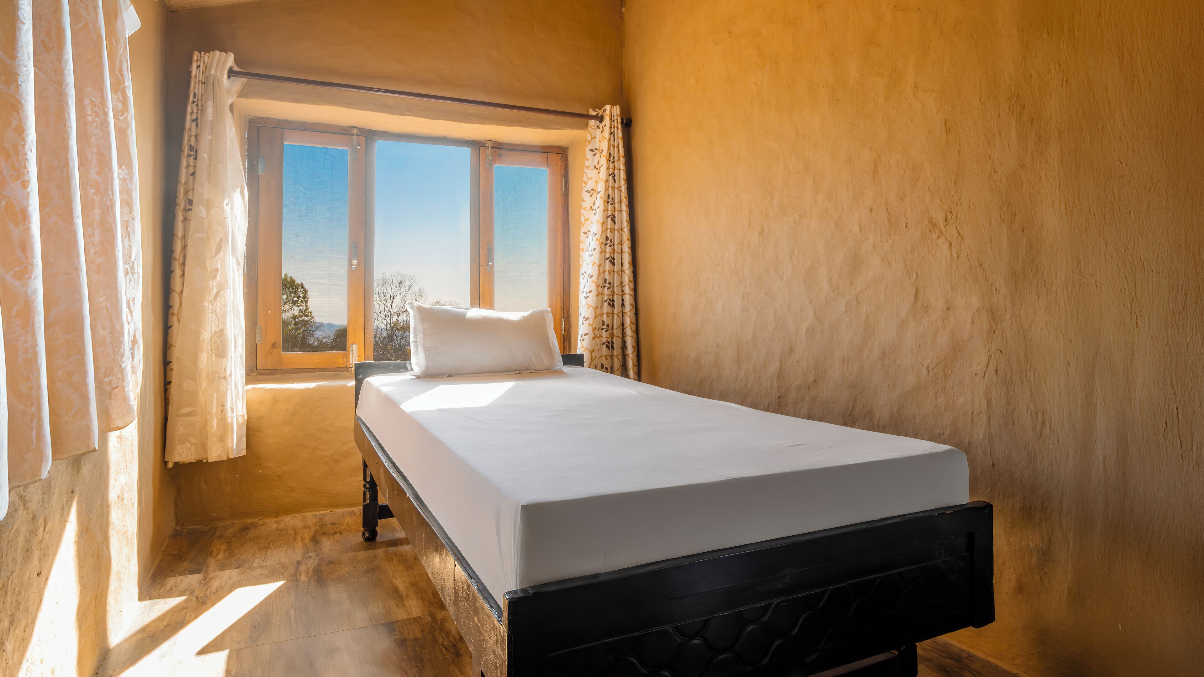 A single bed in a room with a large window, wooden flooring, and a plain wall.