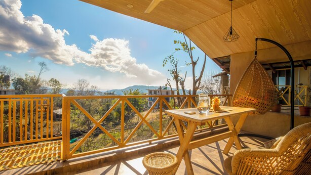A balcony with a wooden railing, a hanging chair, a table with a glass, and a view of trees and mountains. - Dormigo  (Baijnath)