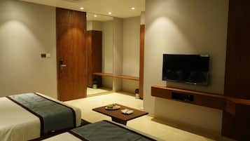 Quarto de hotel com uma cama, um sofá, uma televisão fixada na parede, uma mesa pequena com uma travessa de fruta e um guarda-roupa de madeira.