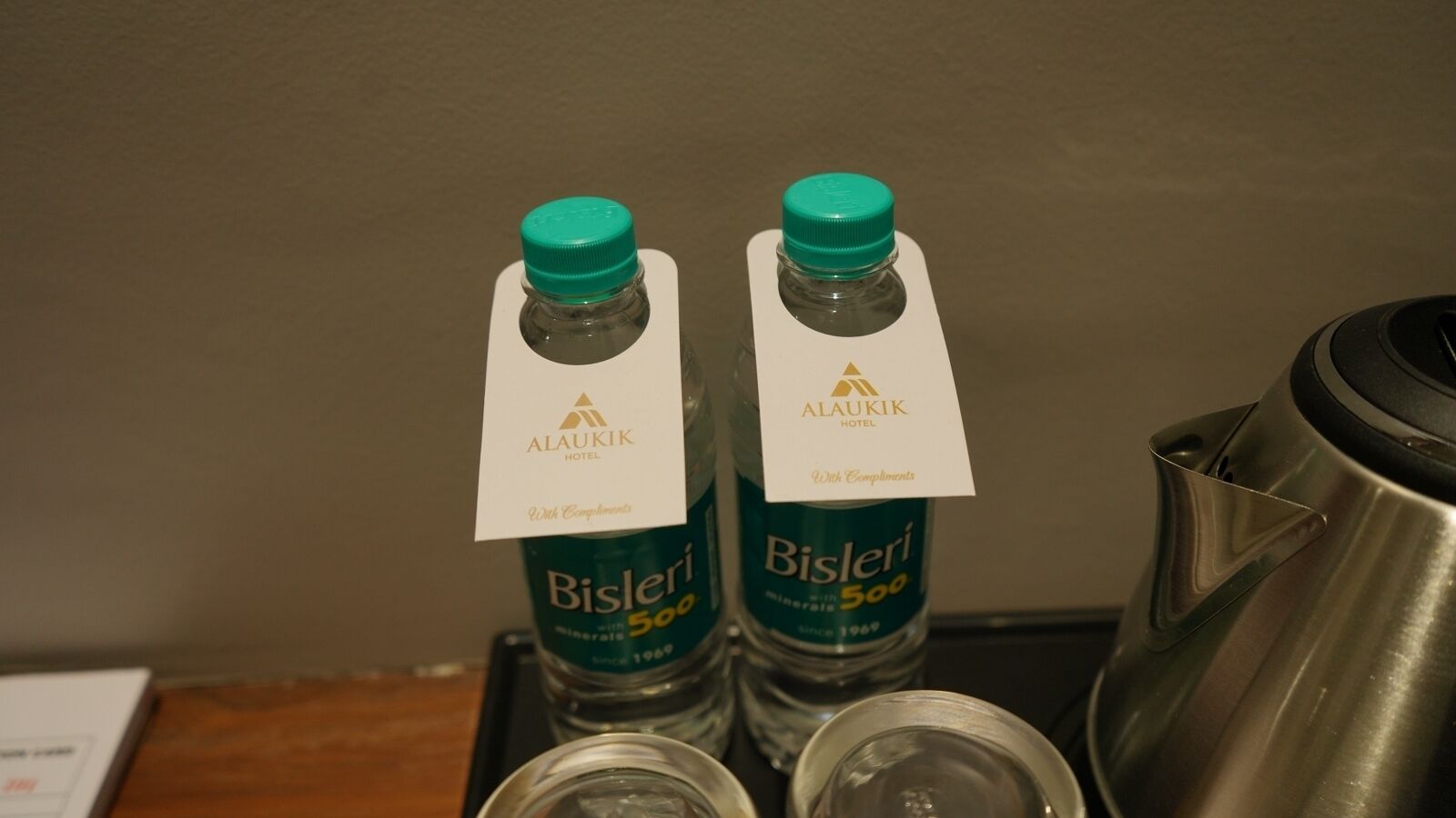 Two bottles of Bisleri 500 mineral water with Alaukik Hotel branding, placed on a tray next to a stainless steel kettle.