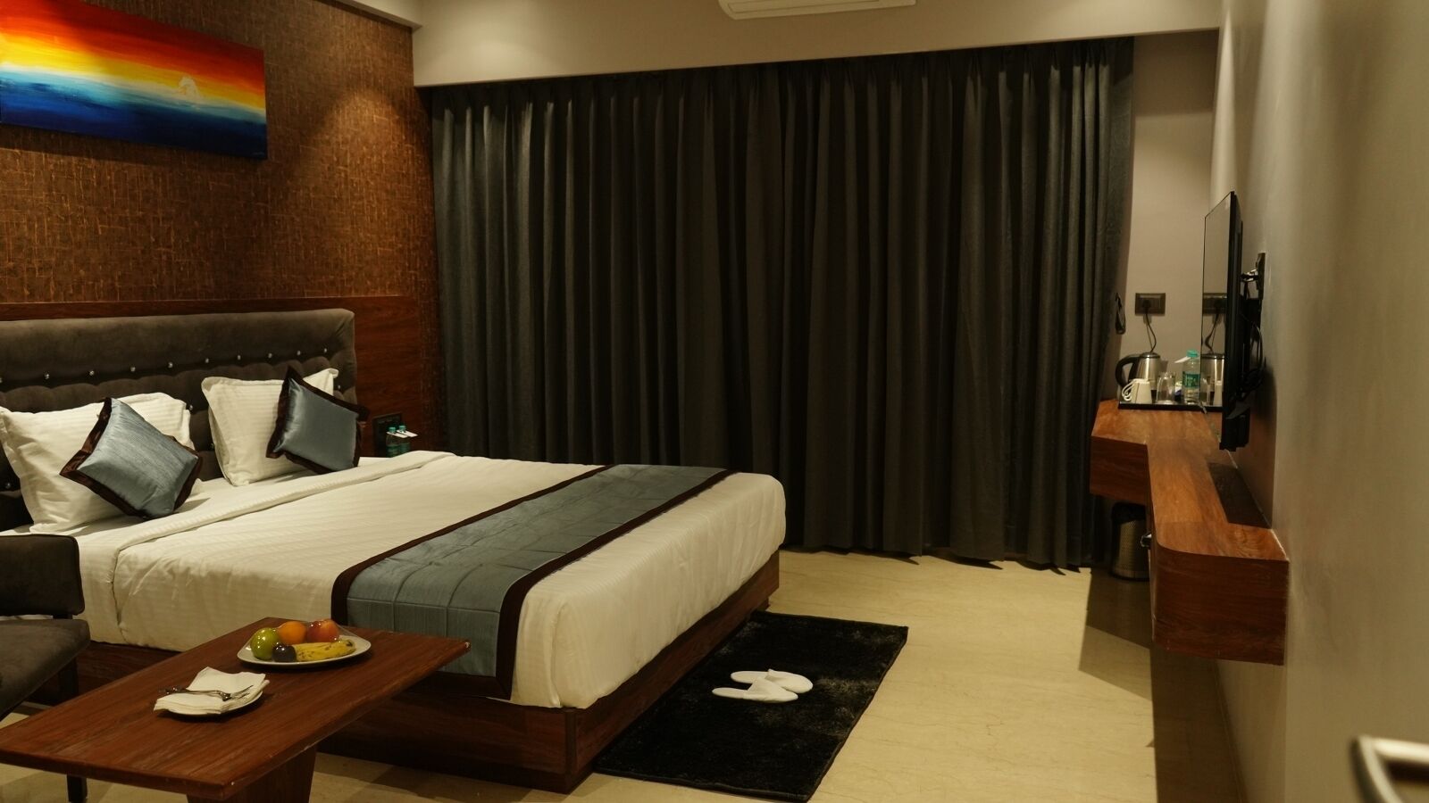 A hotel room with a large bed, a wooden headboard, a small table with fruit, a black rug, and a wooden side table with toiletries.