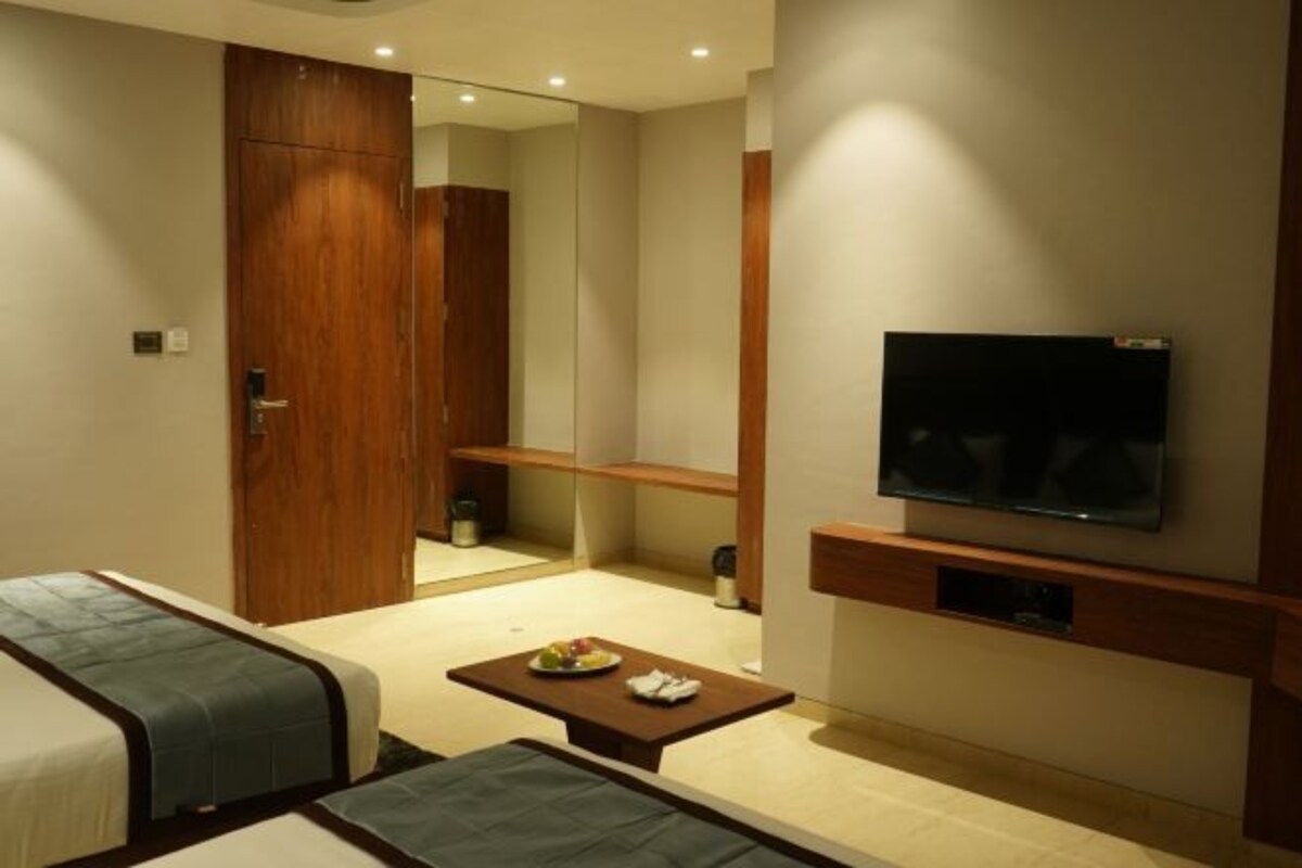 A hotel room with a bed, a TV mounted on the wall, a small table with a plate of fruit, and a mirror.