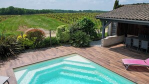A swimming pool with a wooden deck, surrounded by lush greenery and a vineyard in the background. - Heated pool, House in the heart of the vineyards (Puisseguin)