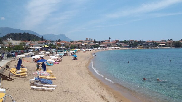 A beach with sun loungers and umbrellas, a clear blue sea, and buildings in the background.