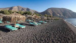 A beach with lounge chairs and thatched umbrellas, a rocky shoreline, and mountains in the background. - Amphora Helios Gem - Private Jacuzzi and Views (Santorini)