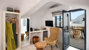 A modern room with a wardrobe, a wicker chair, a small round table, a flat-screen TV, and a large window. - Amphora Helios Gem - Private Jacuzzi and Views (Santorini)
