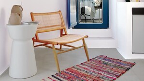Ein kleiner, heller Raum mit einem Korbstuhl, einem weißen Hocker und einem bunten Webteppich. Ein großes Fenster mit blauem Rahmen bietet einen Blick ins Freie.