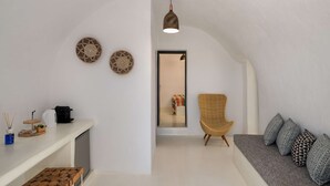 A modern interior with a white wall, a wooden bench, a grey sofa with patterned cushions, a wicker chair, and a hanging pendant light. - Amphora Selene Seaview Getaway in Akrotiri (Santorini)