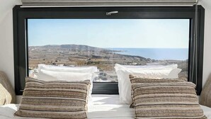 A bedroom with a large window offering a view of a coastal landscape. - Amphora Thalassa Seaview Gem w Private Jacuzzi (Santorini)