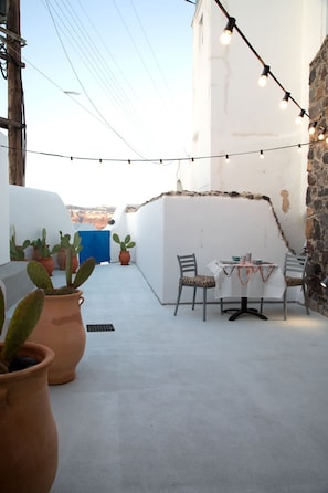 A rooftop terrace with a table and chairs, potted cacti, string lights, and a view of the landscape. - Amphora Charm Suite - Akrotiri Escape (Santorini)