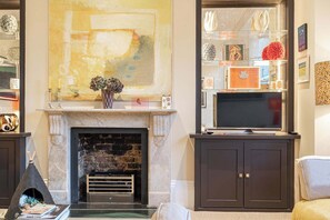 A living room with a fireplace, a television, and a bookshelf with various decorative items. - Lovely 1 Bedroom Flat in Chelsea (London)
