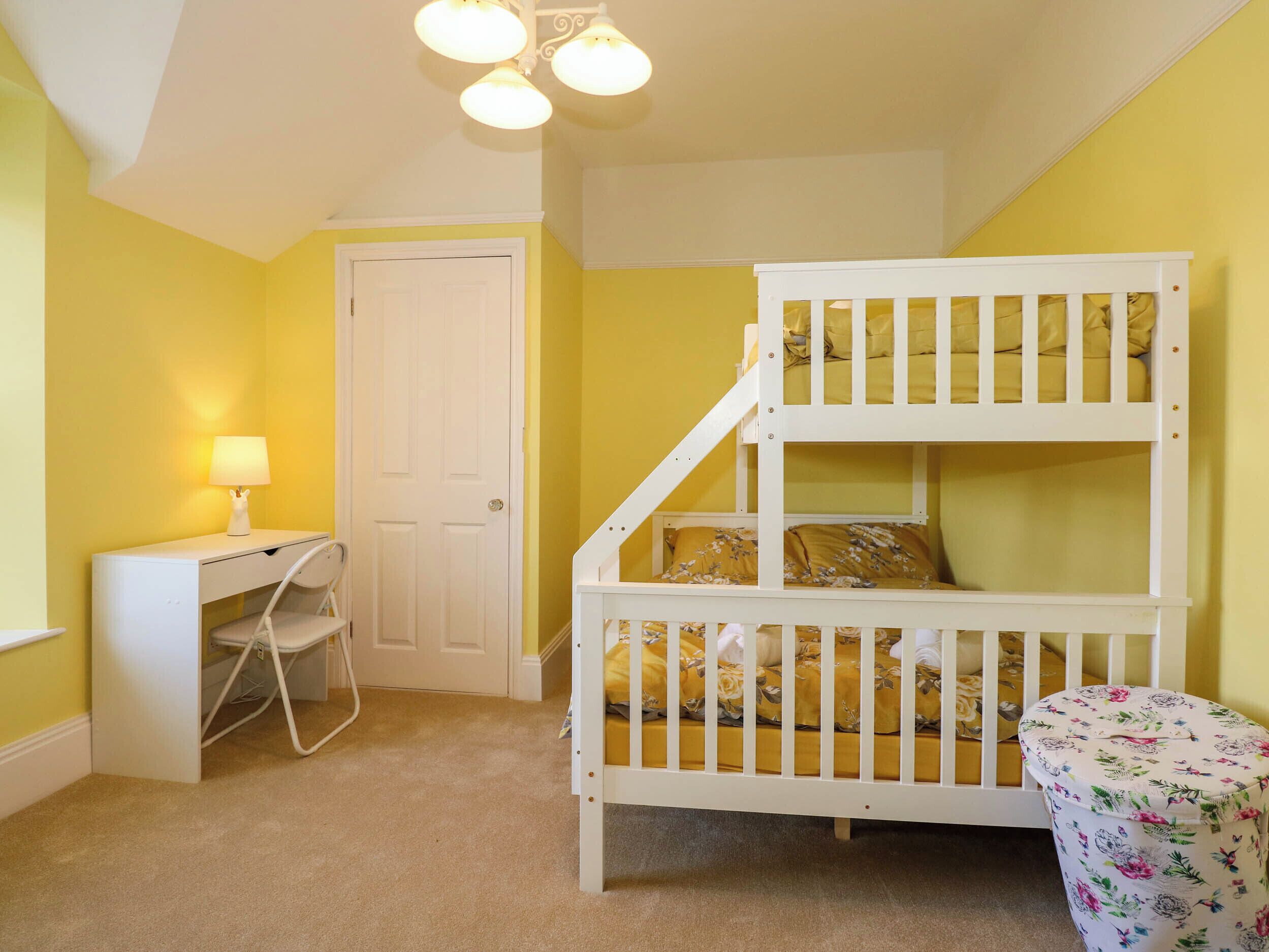 Una cama litera para niños con escritorio y silla, una lámpara y un otoman floral en una habitación con paredes amarillas y una puerta blanca.