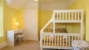Una cama litera para niños con escritorio y silla, una lámpara y un otoman floral en una habitación con paredes amarillas y una puerta blanca.