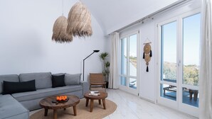 A modern living room with a grey sofa, two wooden coffee tables, a woven rug, and a large window with a view of the outdoors. - Zen Villas (Akrotiri)