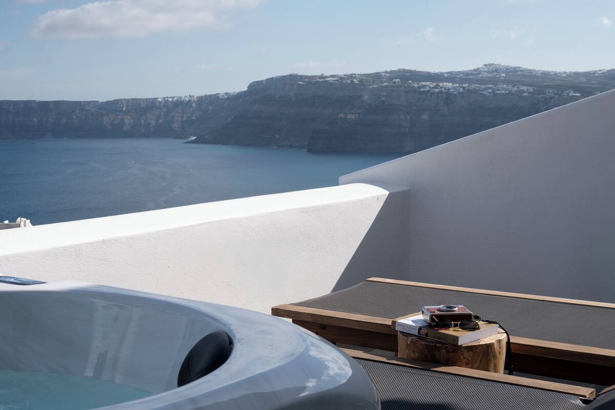 A sunlit balcony with a hot tub, lounge chairs, and a view of the sea and cliffs.