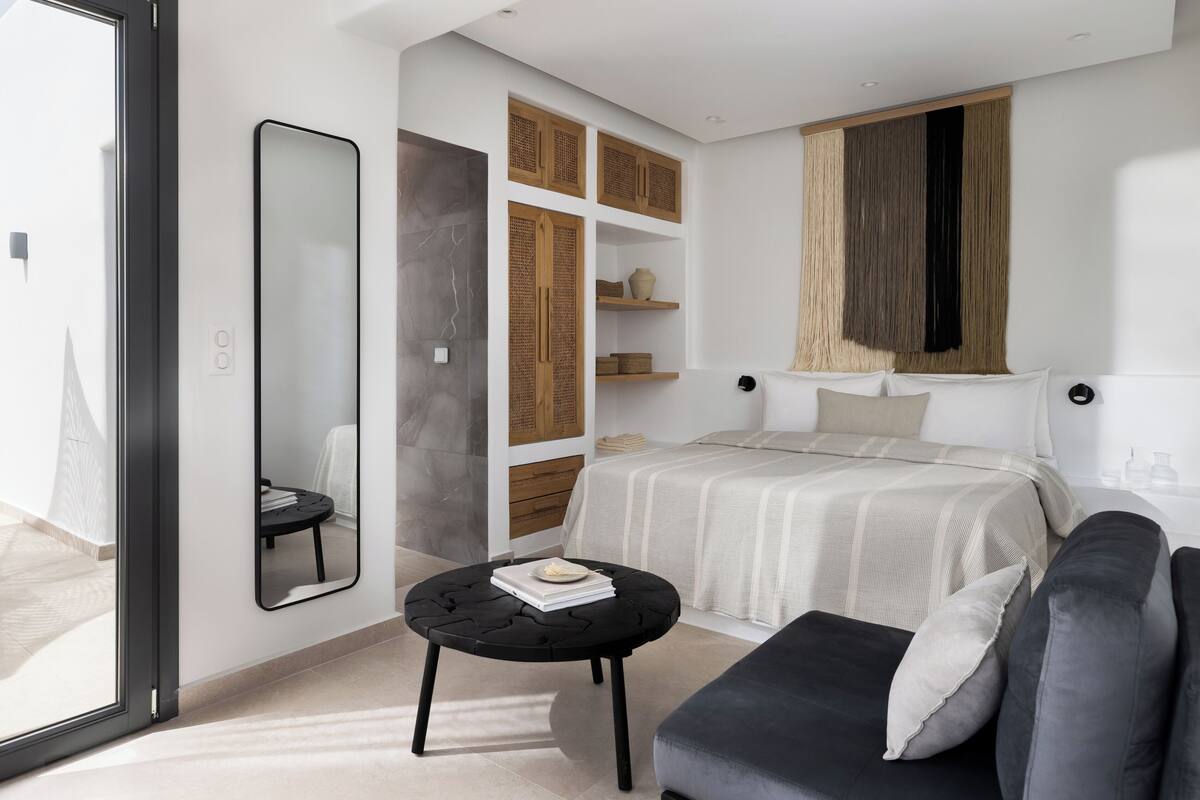A modern bedroom with a bed, a sofa, a round black table, a large mirror, and built-in wooden shelves.