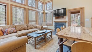 Un salón con sofá, mesa de centro y mesa de comedor. Hay ventanas amplias, una chimenea y un televisor montado en la pared.