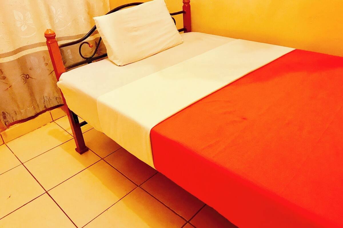 Una cama individual con colchón blanco y manta roja, cabecero de madera y almohada blanca.