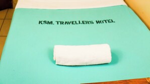 A single bed with a white bedspread and a white towel folded at the foot. The bed is in a room with yellow walls and a wooden headboard. The text "KSM. TRAVELLERS HOTEL" is visible on the bedspread. - Kisumu Travelers Hotel (Kisumu)