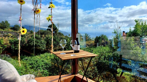 Ein Tisch mit zwei Weingläsern und einer Flasche, vor einer malerischen Landschaft mit viel Grün und blauem Himmel.