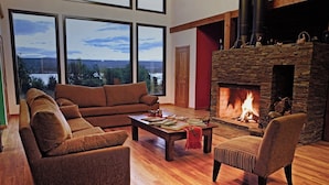 A cozy living room with a stone fireplace, a brown sofa, a coffee table with books, and a view of a lake through large windows. - Lago Rosario Lodge (Trevelin)