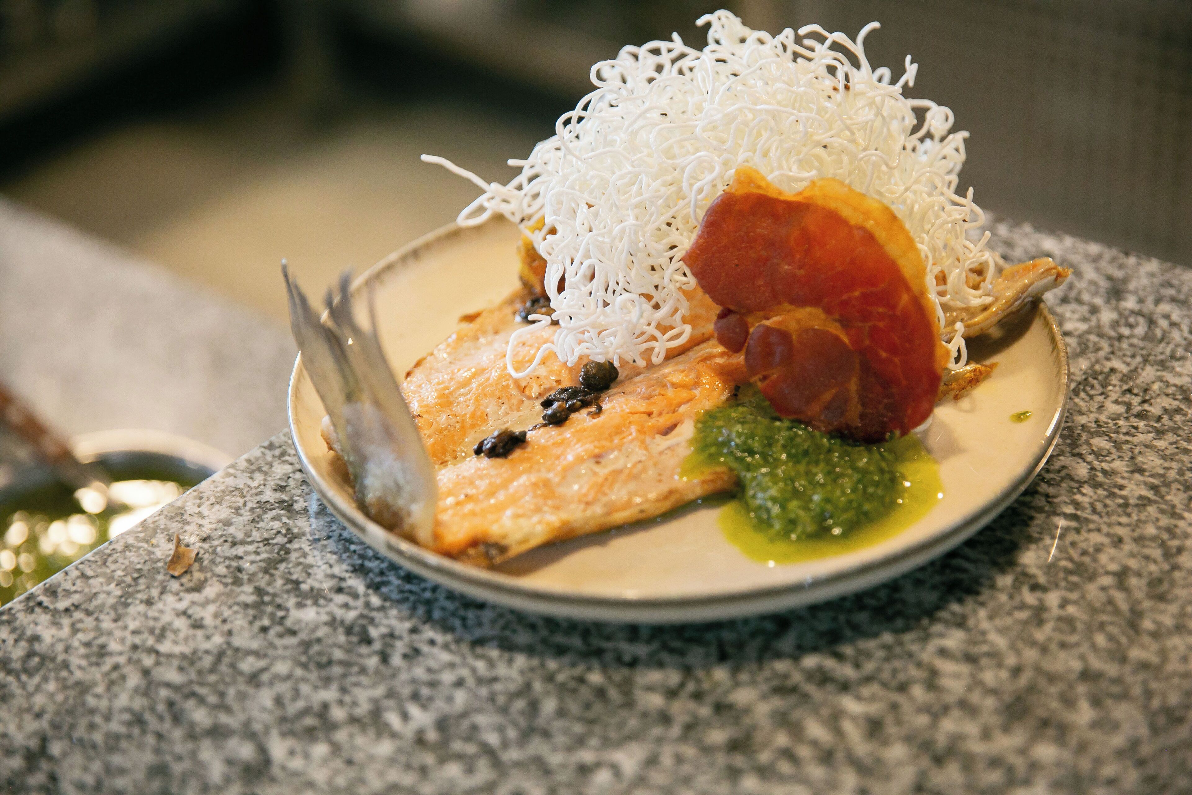 A plate with salmon, noodles, a slice of red meat, and a green sauce.
