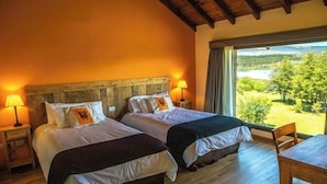 A room with two beds, wooden headboard, bedside lamps, a wooden table, and a view of a landscape through a large window. - Lago Rosario Lodge (Trevelin)