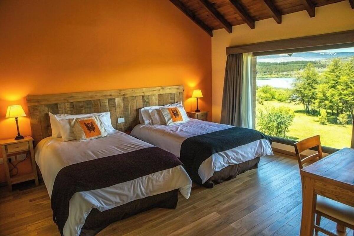 A room with two beds, wooden headboard, bedside lamps, a wooden table, and a view of a landscape through a large window.