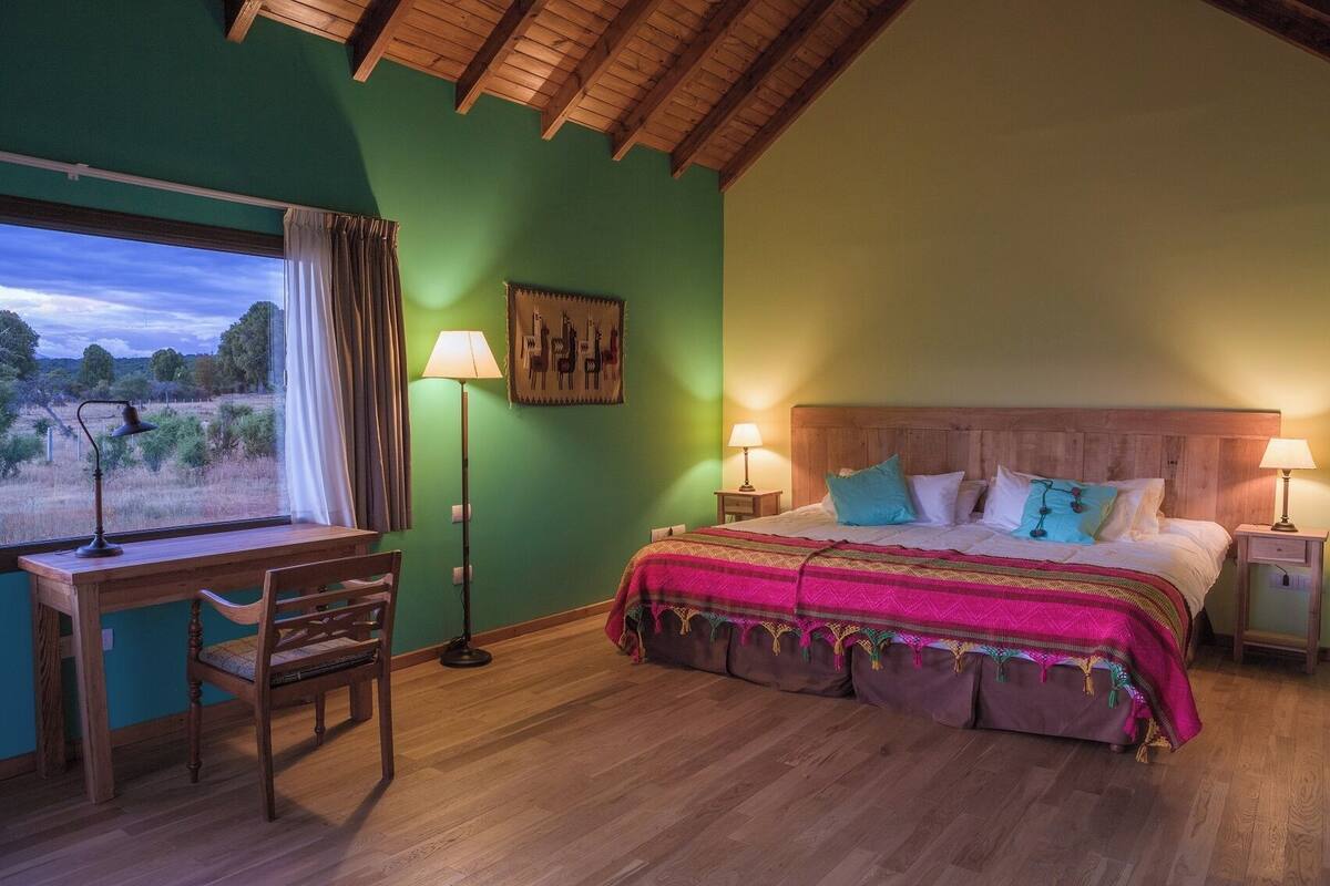 A bedroom with a wooden desk, a bed with colorful bedding, a nightstand, and a lamp.